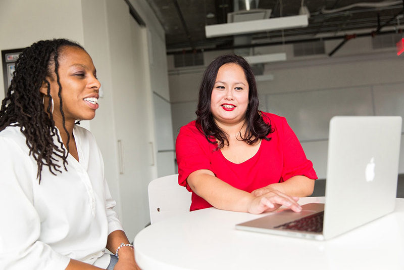 two women smiling and talking together in front of a laptop computer