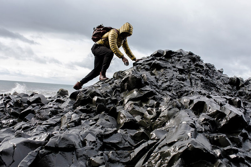 man wearing a backpack in trousers, leather shoes, and puffer coat climbing a rocky section of shoreline by the ocean